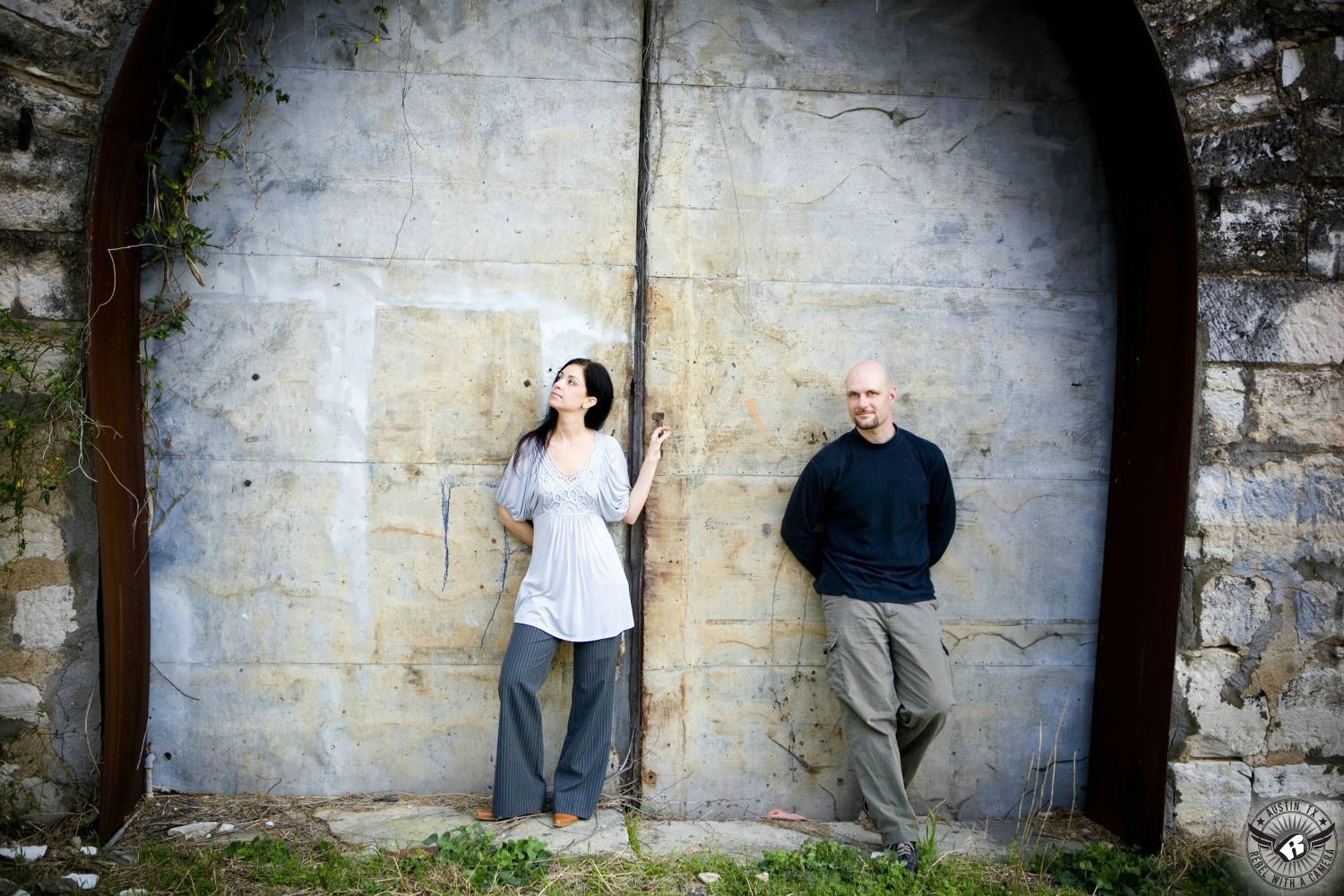 Dark haired Italian lady wearing a grey pleated, low cut thigh length blouse with grey dress pants stands leaning in a rusted metal arch against a galvanized metal shop door next to a guy wearing a black long sleeve shirt  and grey pants at a vacant old building in Taylor in this tense engagement image in rural Austin. 